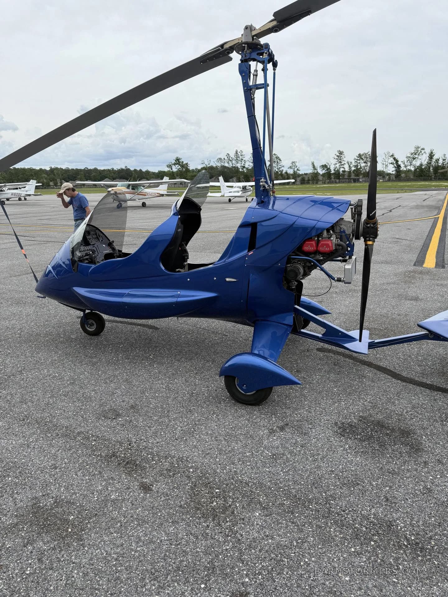 A blue autogyro is parked on an asphalt tarmac, viewed from the right side, with a person and other aircraft in the background.