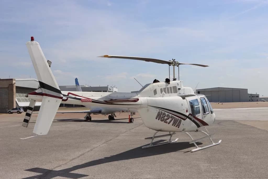 A white Bell 206 helicopter with N827WA registration on the side, parked on an airfield with other aircraft in the background.