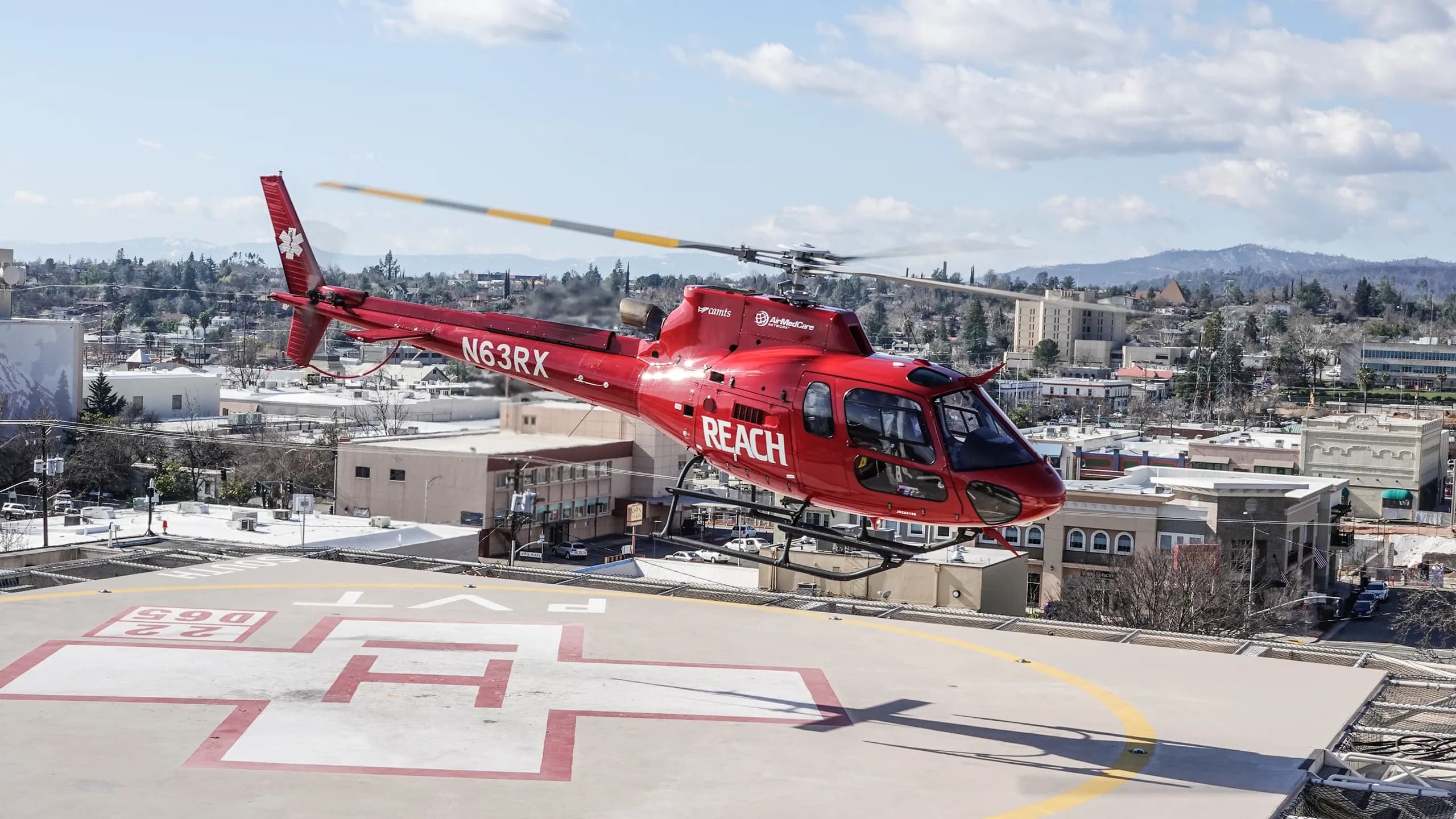 A red REACH air ambulance helicopter takes off from a helipad in a city.