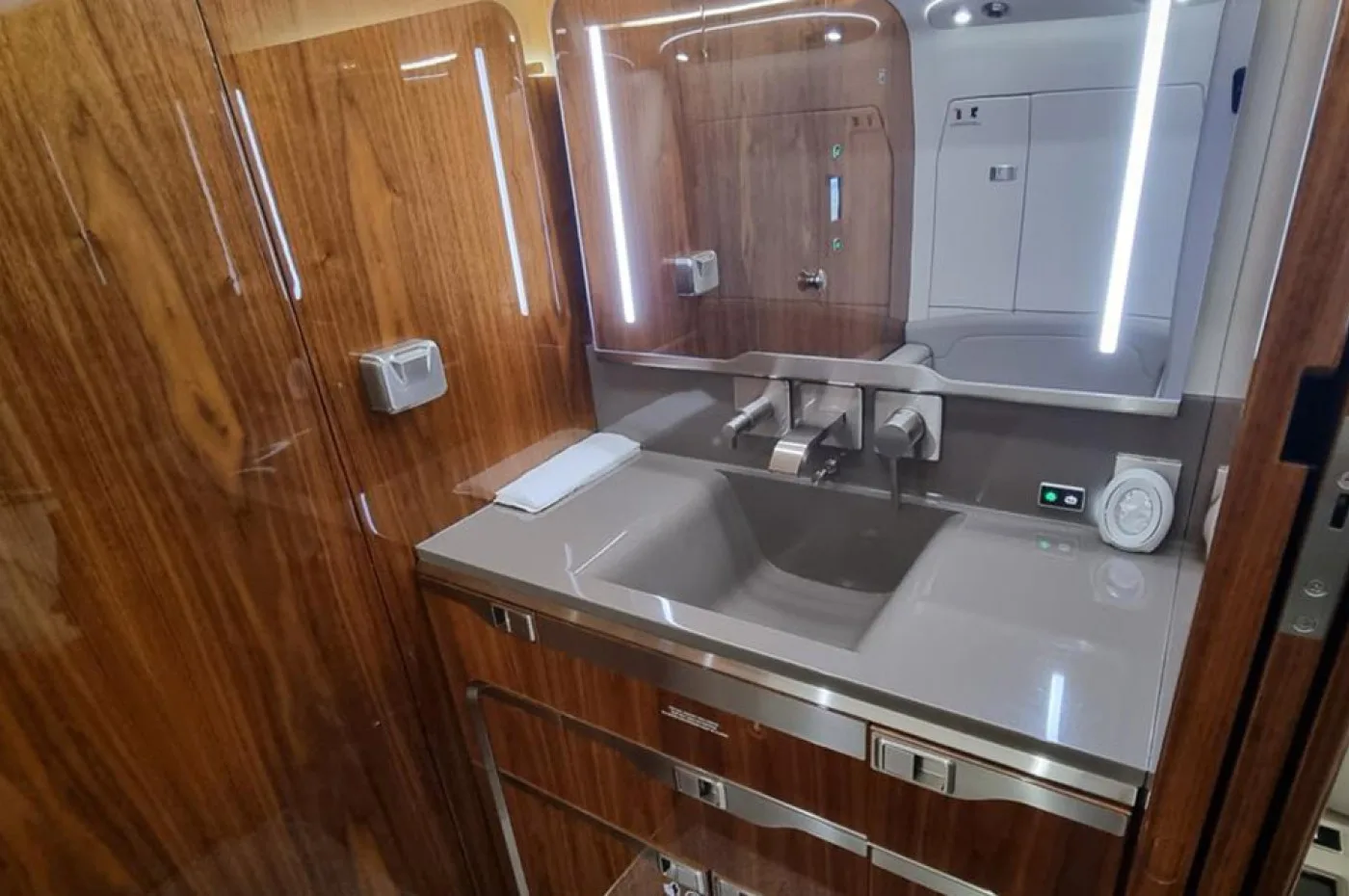 A modern aircraft lavatory features a grey sink, chrome faucet, an illuminated mirror, and polished wood paneling.