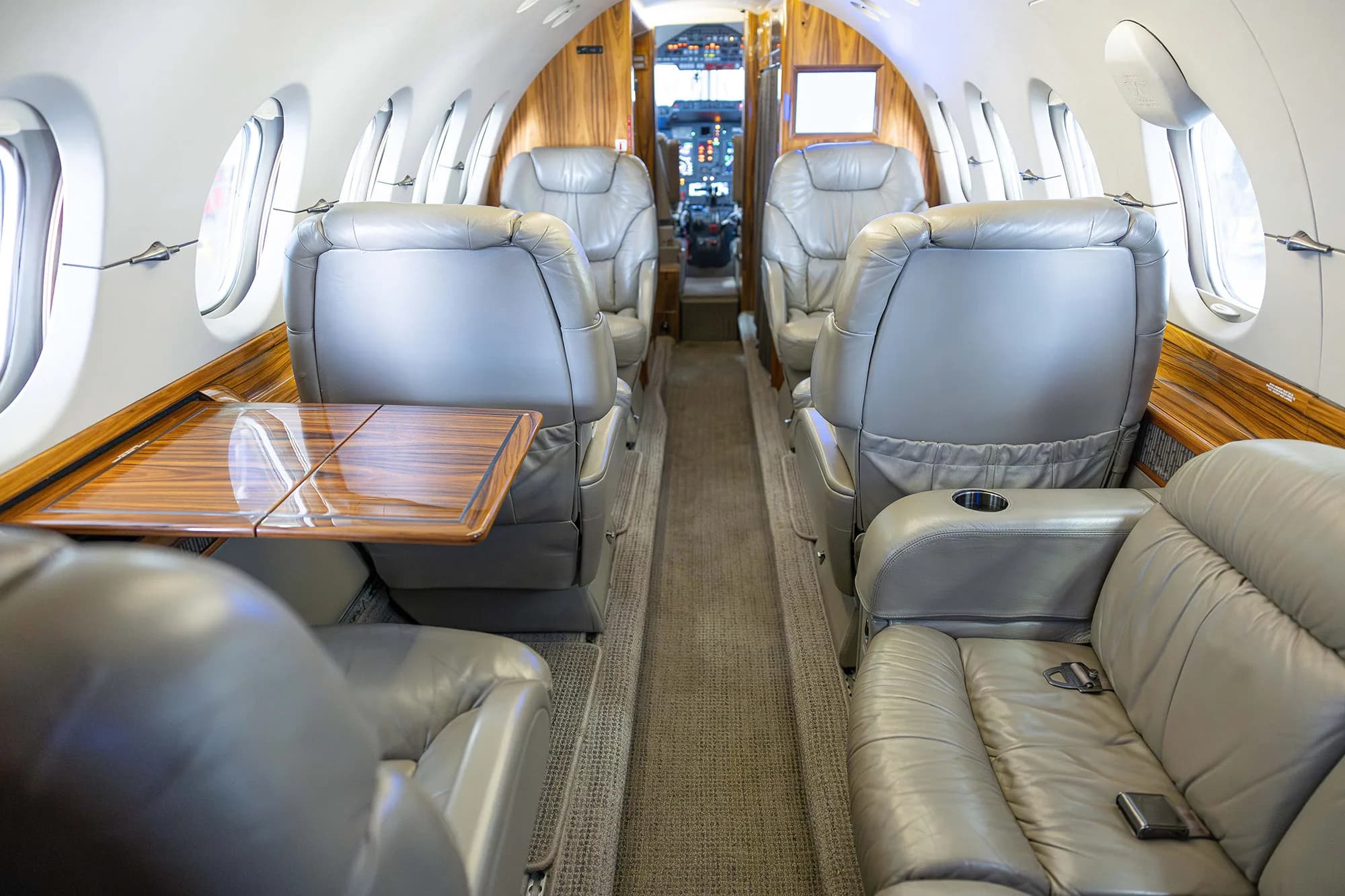 The interior cabin of a business jet with grey leather seats, a side-facing couch, and wood paneling.
