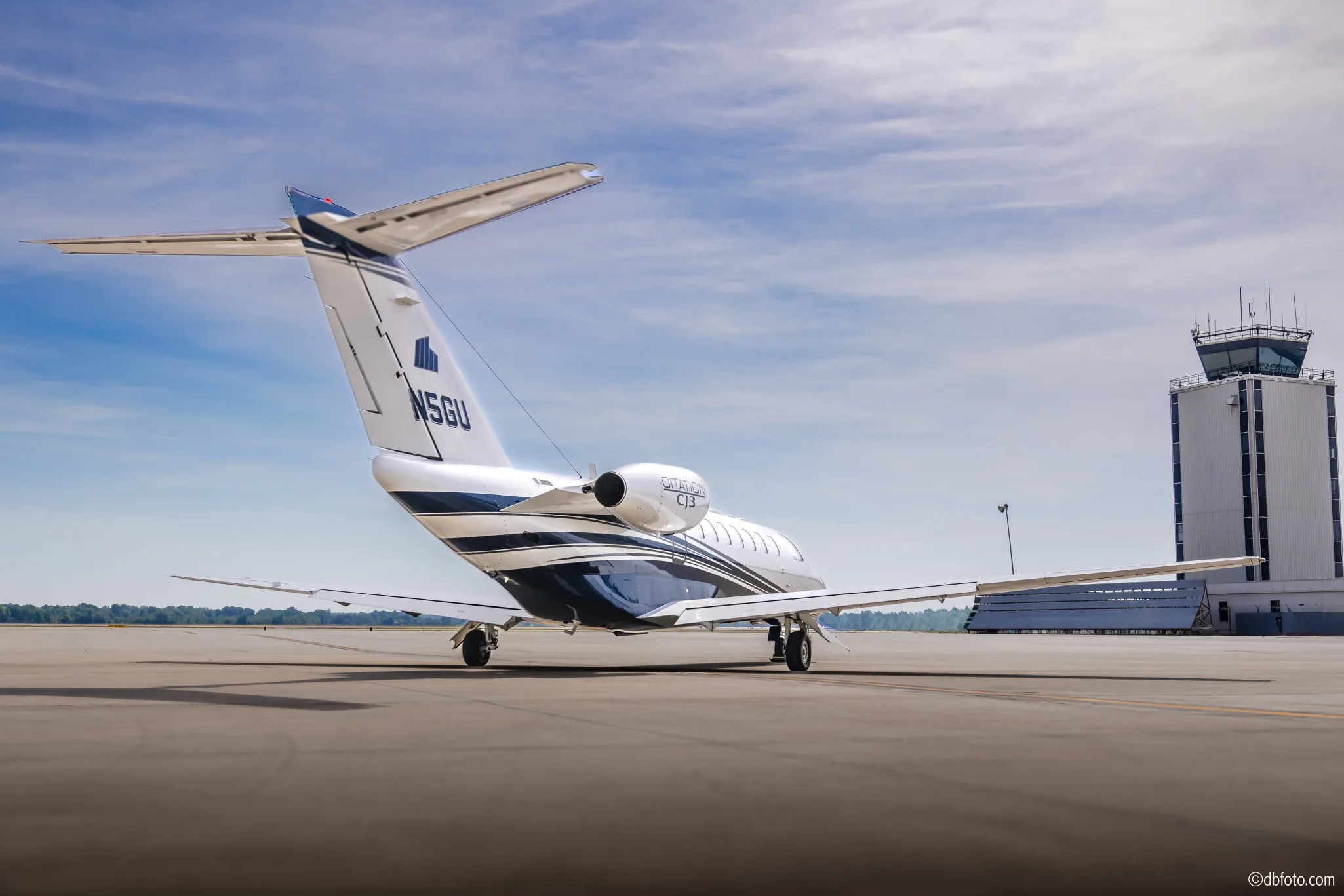 A rear-side view of a white Cessna Citation CJ3 business jet with blue and grey stripes, registration N5GU, parked on an airport tarmac with a control tower in the background.