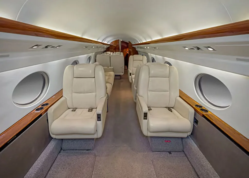 The interior cabin of a private jet features two rows of beige leather seats facing each other, with wood trim along the windows.