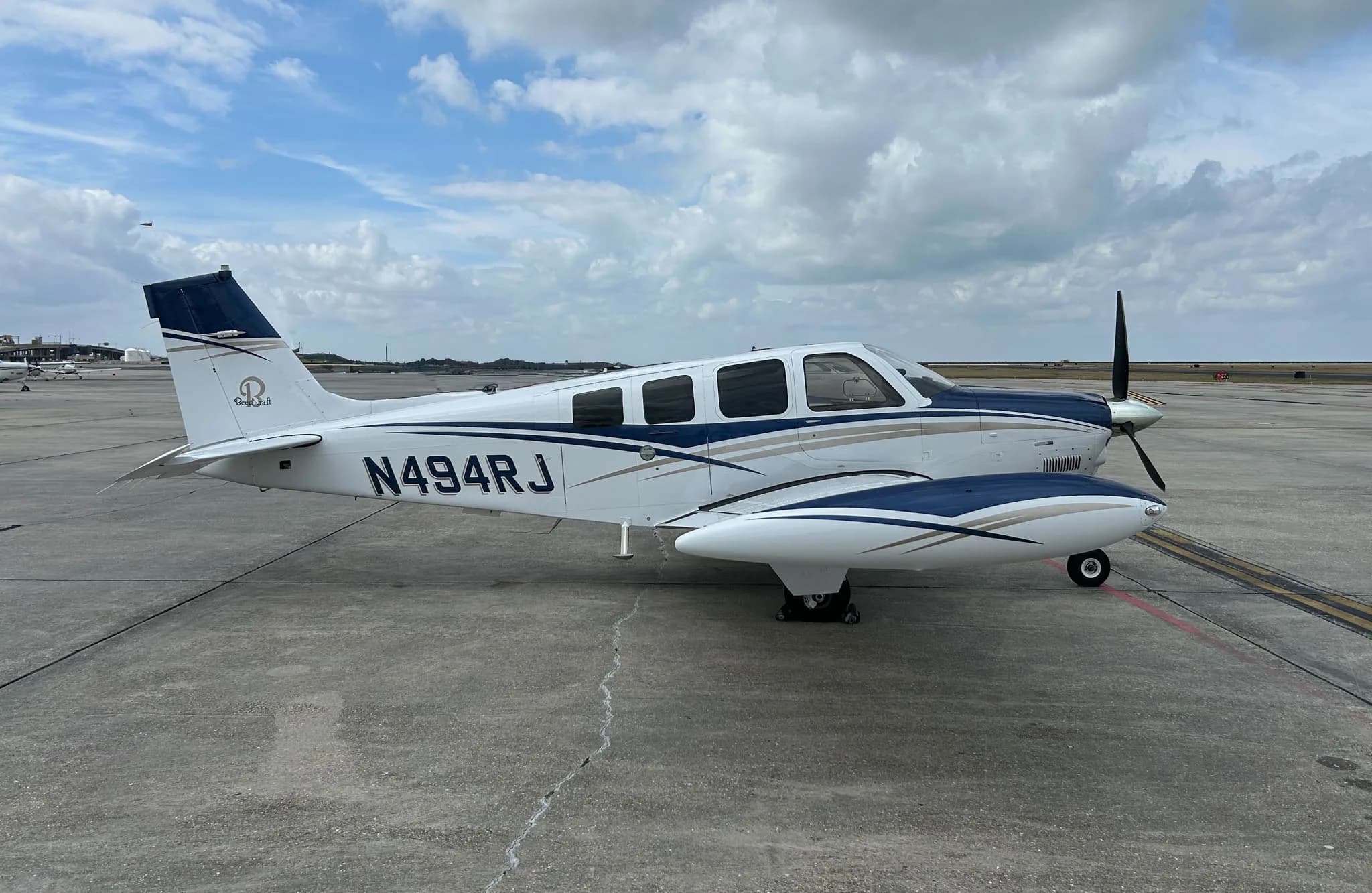 A white and blue Beechcraft Bonanza, N494RJ, is parked on an airport tarmac under a cloudy sky.