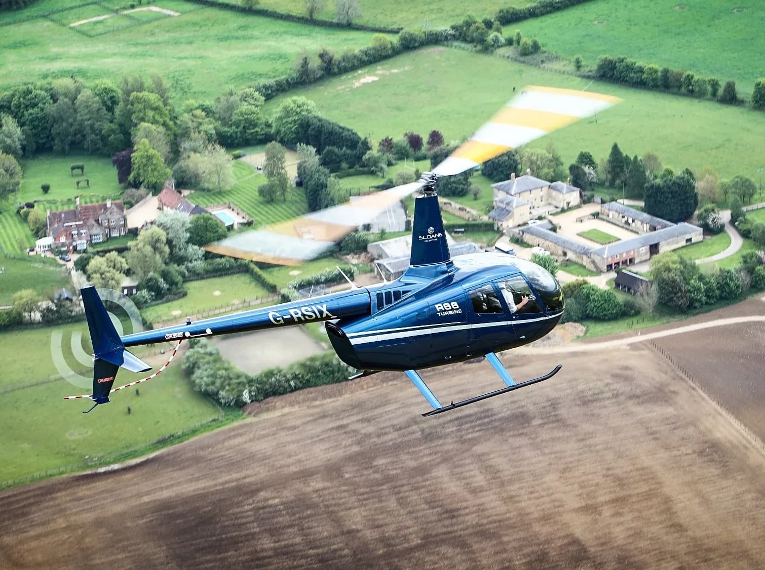 A blue Robinson R66 Turbine helicopter in flight over a rural area.