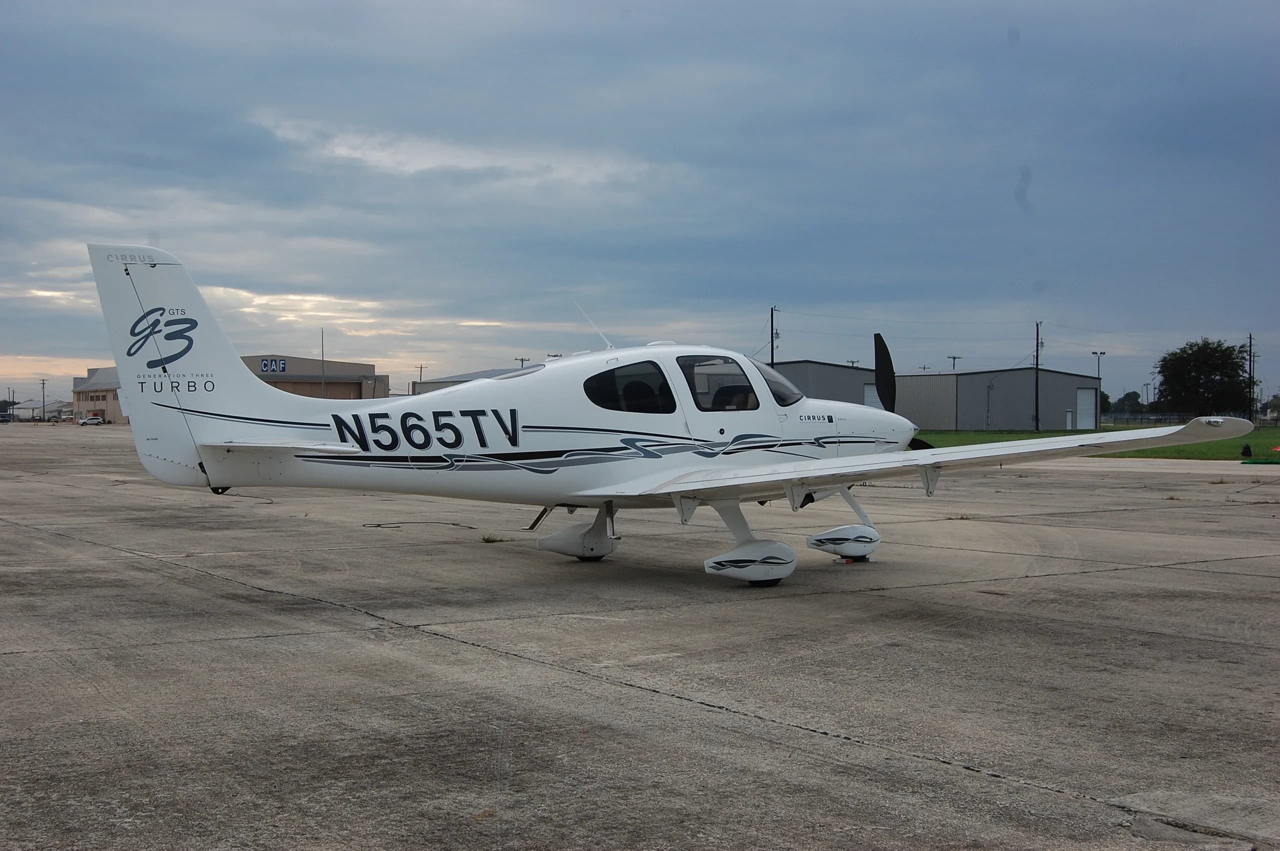 A white Cirrus SR22T aircraft with black and grey stripes, registration N565TV, parked on a concrete tarmac under a cloudy sky.