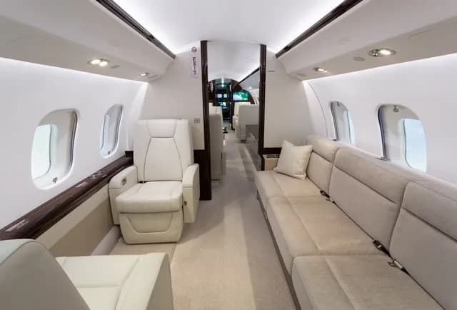 An aircraft cabin with a long beige sofa, individual white seats, and dark wood accents.