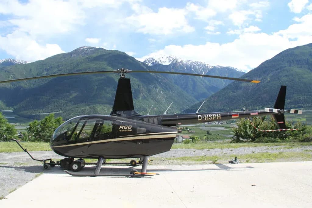 A black Robinson R66 Turbine helicopter with a gold stripe, registration D-HSPH, parked on a concrete pad with green and snow-capped mountains in the background.