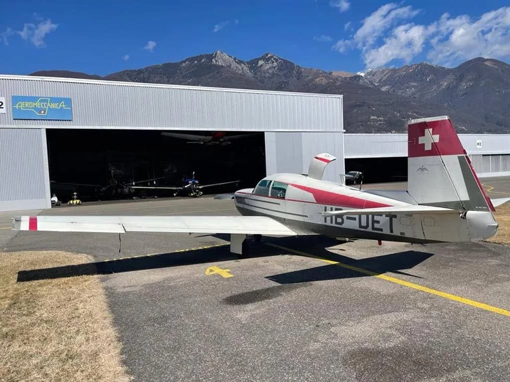A white and red single-engine aircraft, HB-DET, with a Swiss flag on its tail, parked on an airfield next to a hangar.