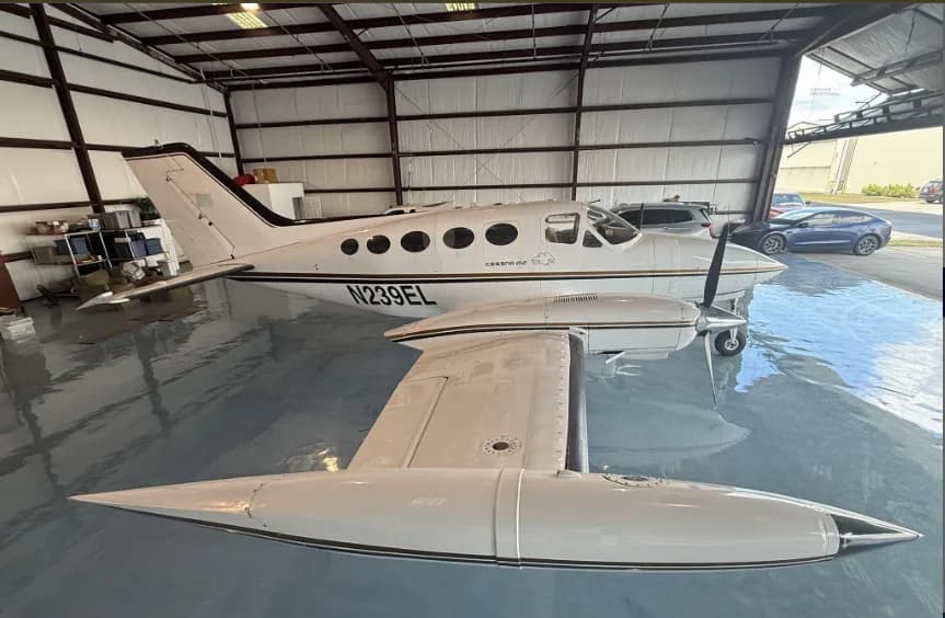 A white Cessna 414 aircraft with gold and black stripes and registration N239EL, parked in a hangar, viewed from the front-right.