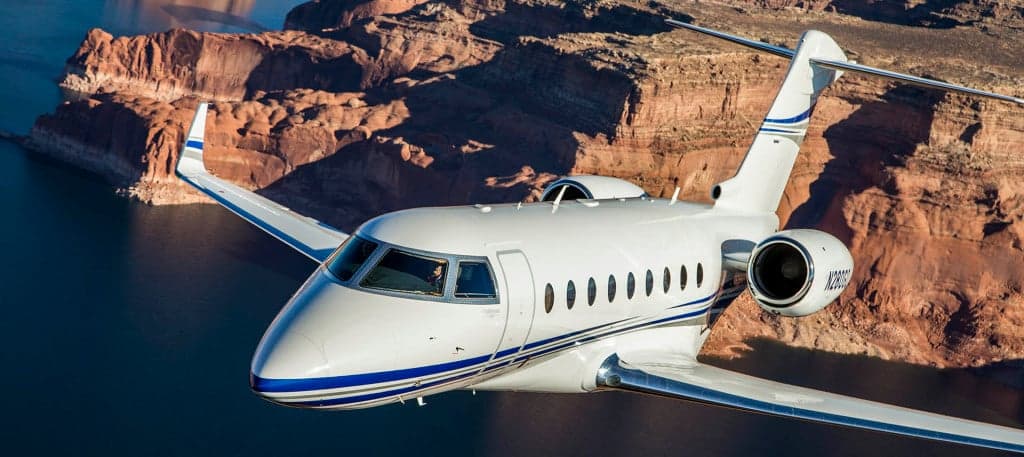 A white private jet flying over a lake and mountains.