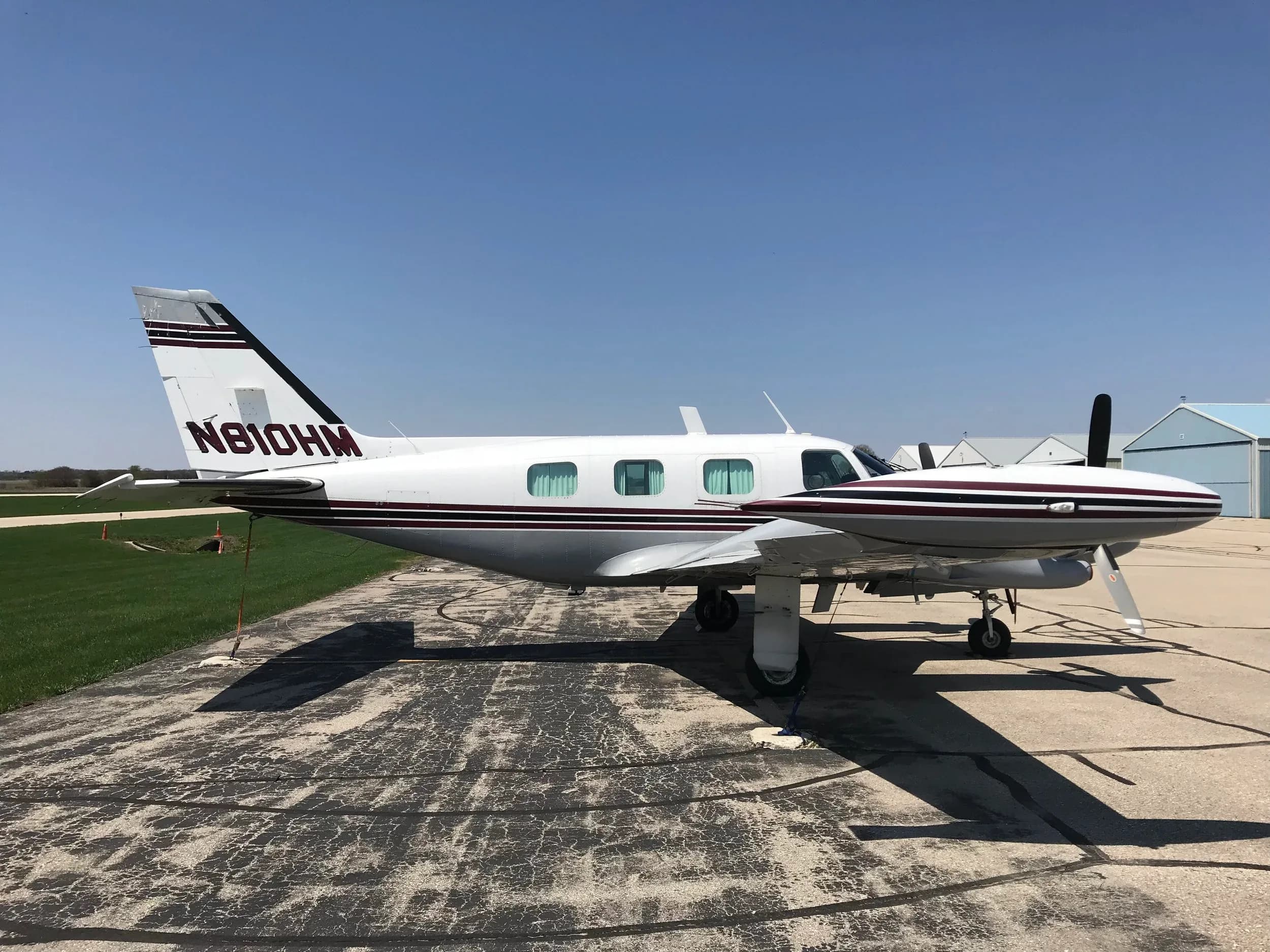 A white twin-engine propeller aircraft with maroon and black stripes, tail number N810HM, parked on an asphalt ramp under a clear blue sky.