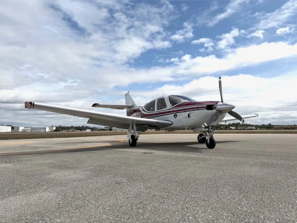 A white and maroon single-engine propeller aircraft is parked on an asphalt tarmac under a partly cloudy sky.