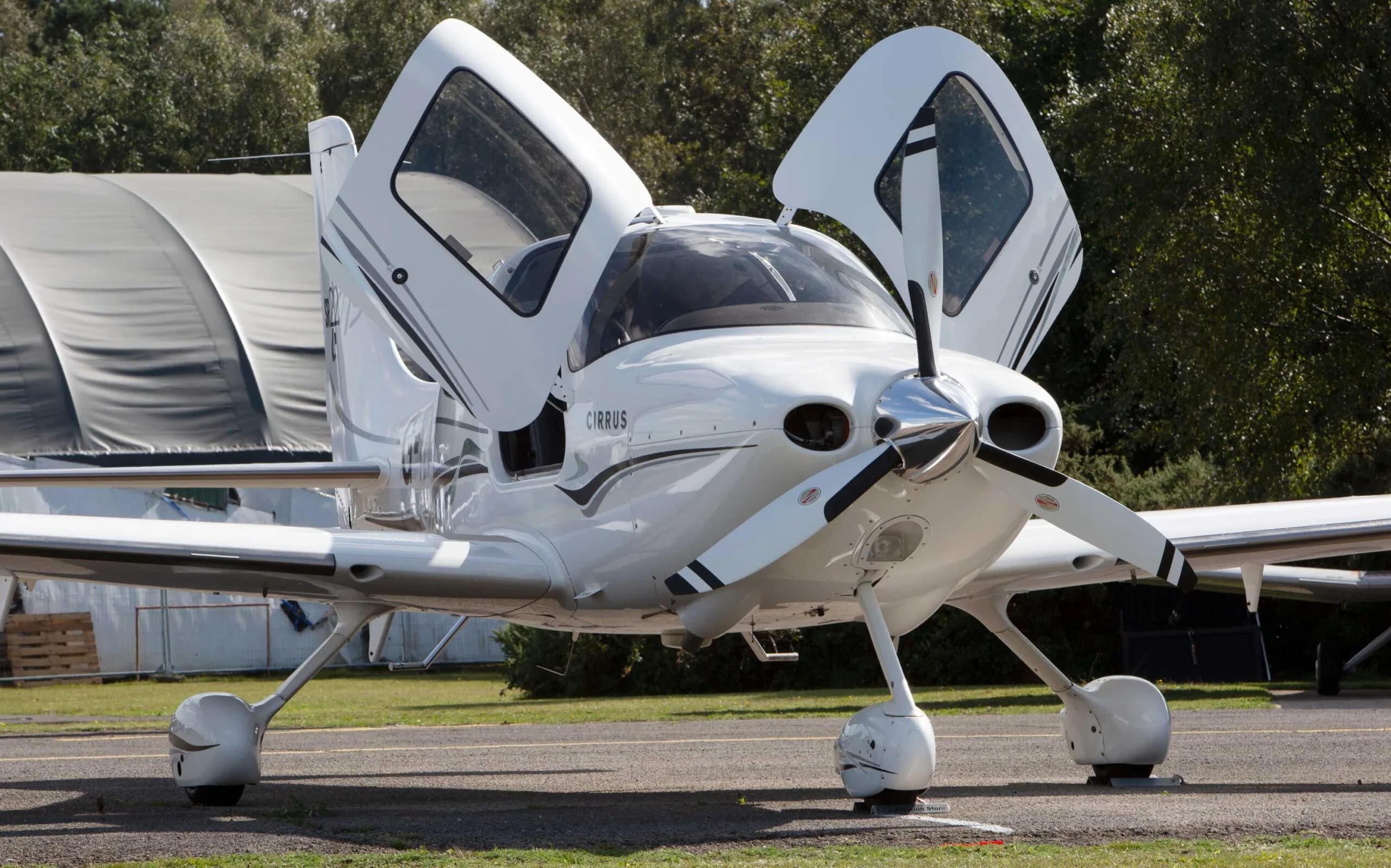 A white Cirrus SR22 TSi aircraft with black and grey stripes, seen from the front-right with its gull-wing doors open on an airfield.