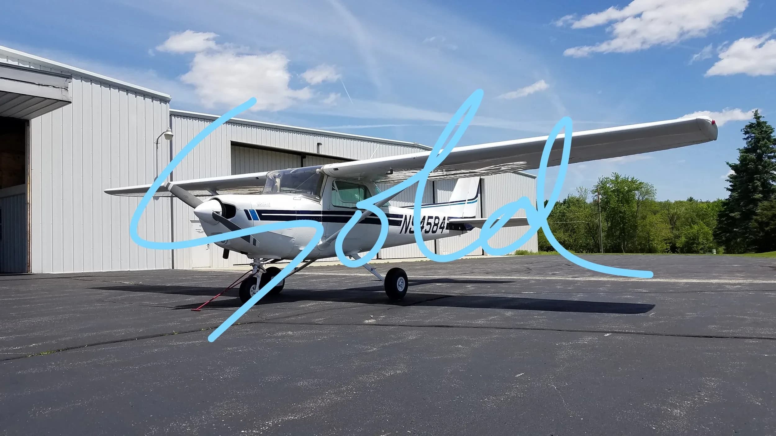 A white Cessna 172 aircraft with blue stripes is parked on an asphalt ramp next to a hangar under a blue sky.