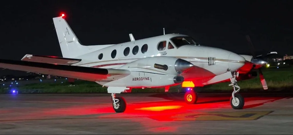 A white Beechcraft King Air C90B with red landing lights illuminated, parked on an airfield at night.