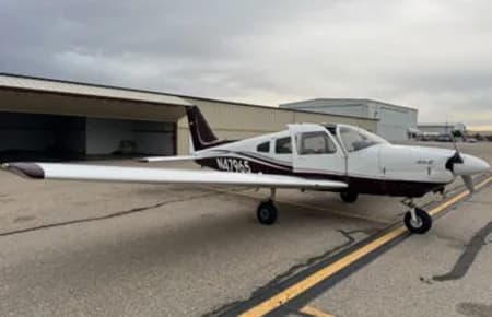 A white and maroon single-engine propeller aircraft, N4796S, parked on an airfield tarmac with hangars in the background under an overcast sky.