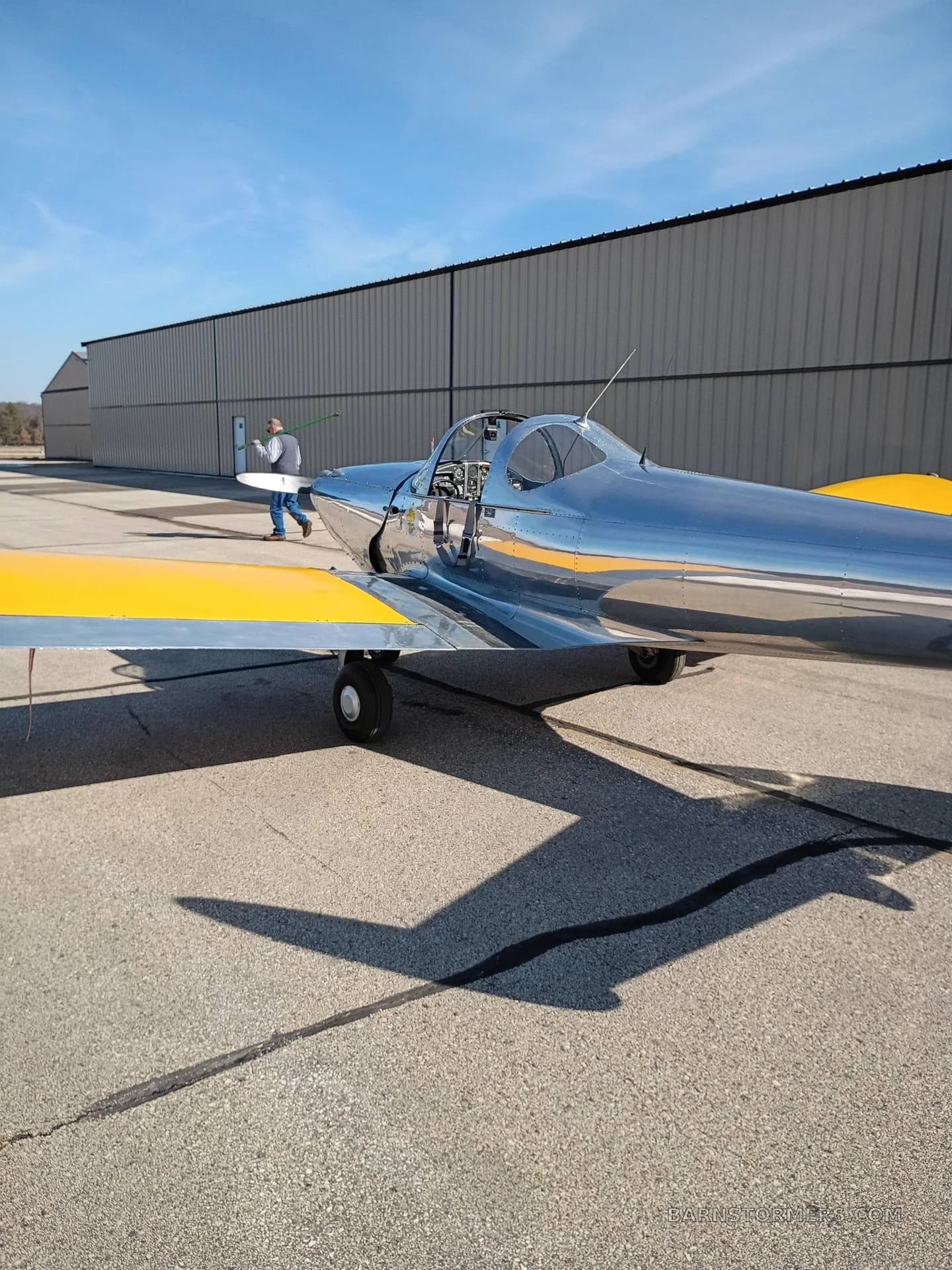 A side view of a polished silver aircraft with yellow wings on an airfield, with a person walking near a hangar in the background.