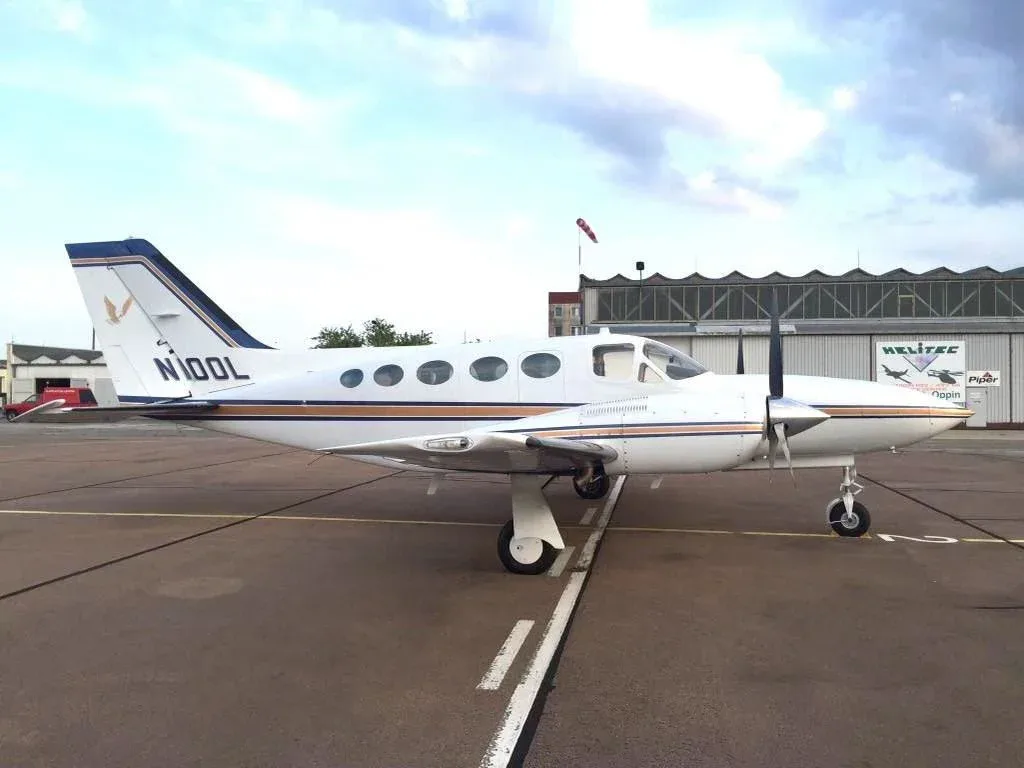 Side view of a white Cessna 421 Golden Eagle aircraft with blue and gold stripes parked on the tarmac next to a hangar.
