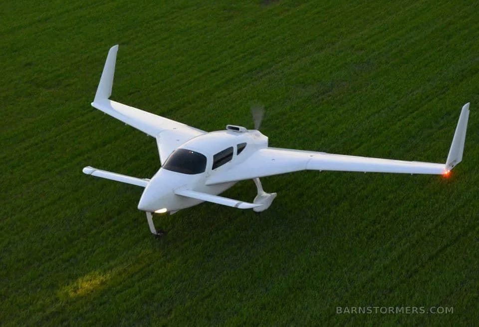 A white, single-engine, pusher-propeller aircraft with twin tail booms flies low over a green field.