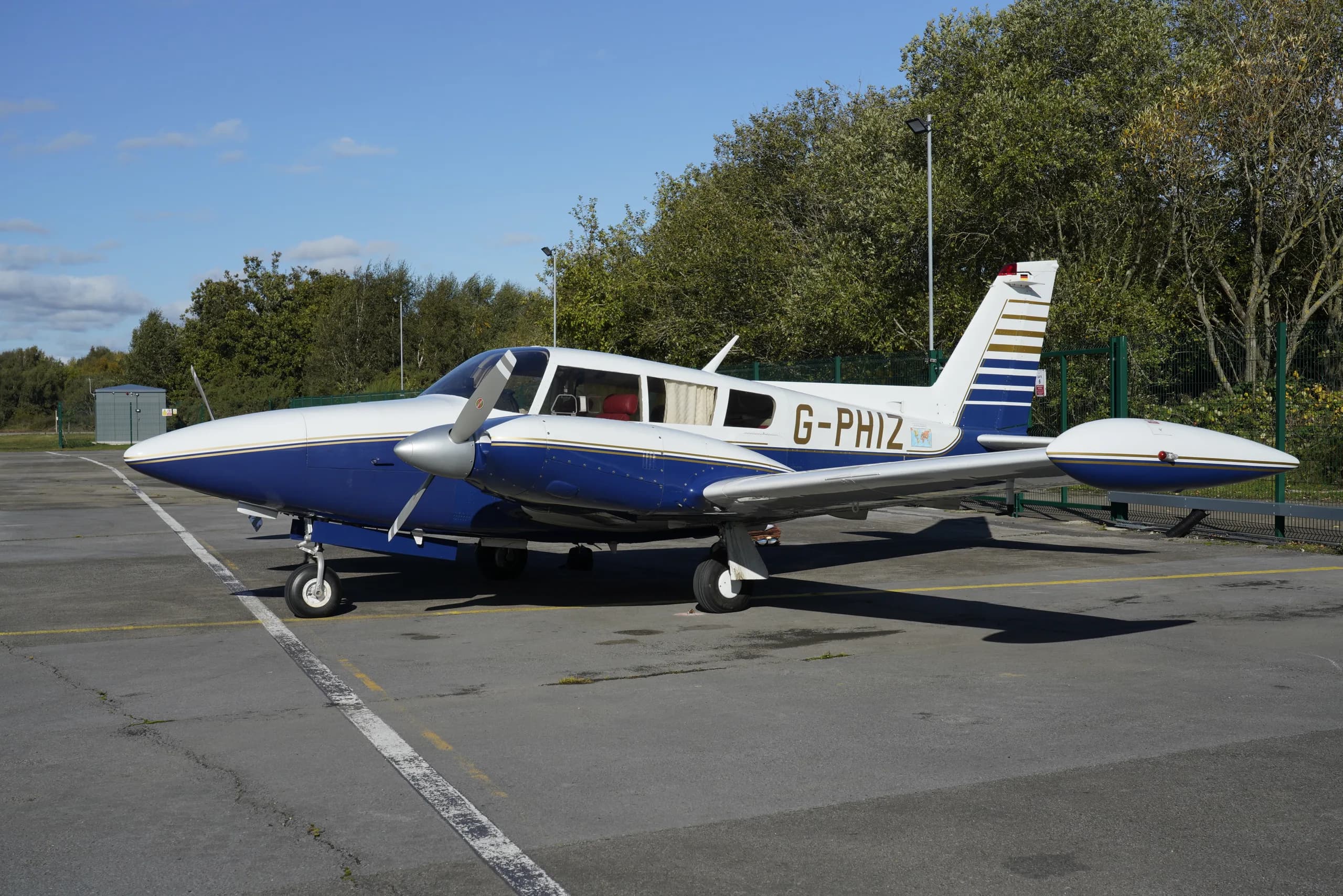 A white and blue single-engine propeller aircraft with registration G-PHIZ parked on an asphalt surface surrounded by trees.