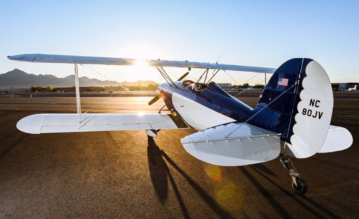 A blue and white biplane, N80JV, is parked on the tarmac with the sun setting in the background.