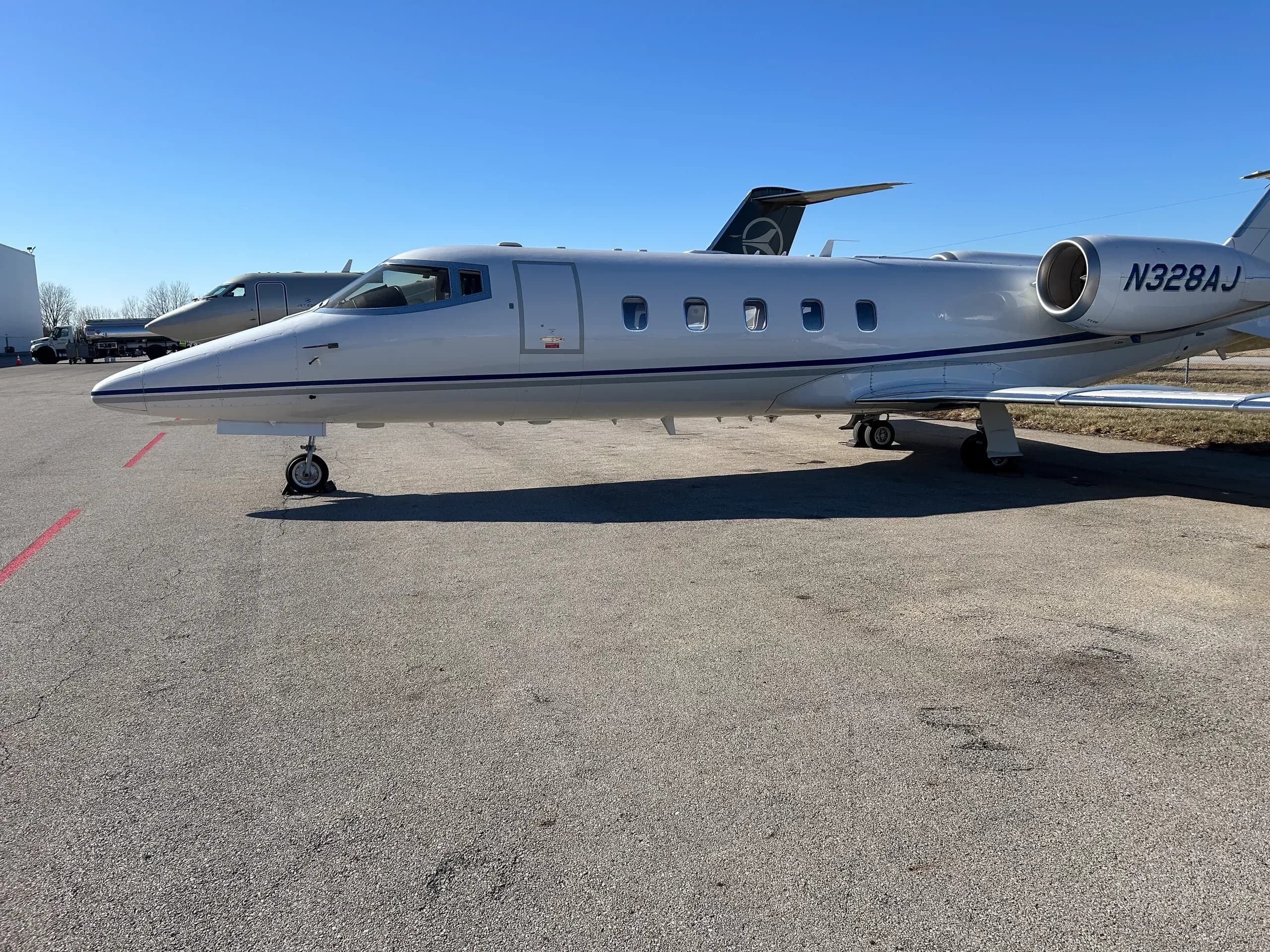 A full side view of the white Learjet N328AJ with blue stripes parked on an airport tarmac.
