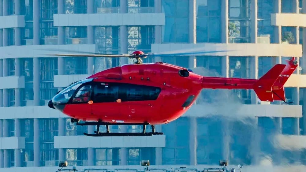A red Airbus H145 helicopter hovers in front of modern glass buildings.