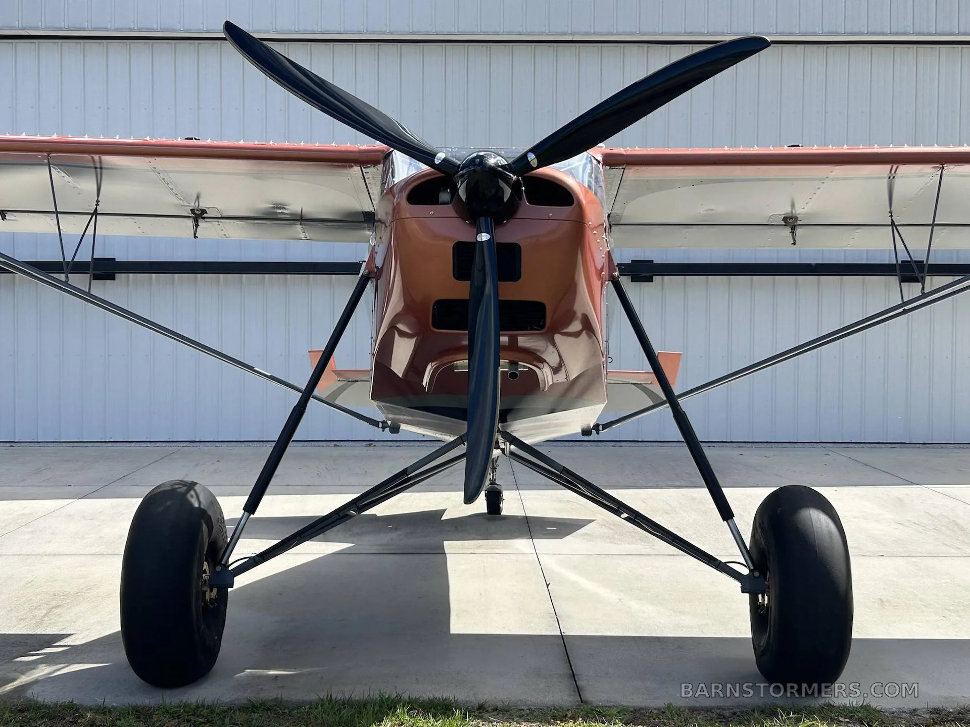 A front view of an orange and grey bush plane with large tires, parked in front of a white hangar.