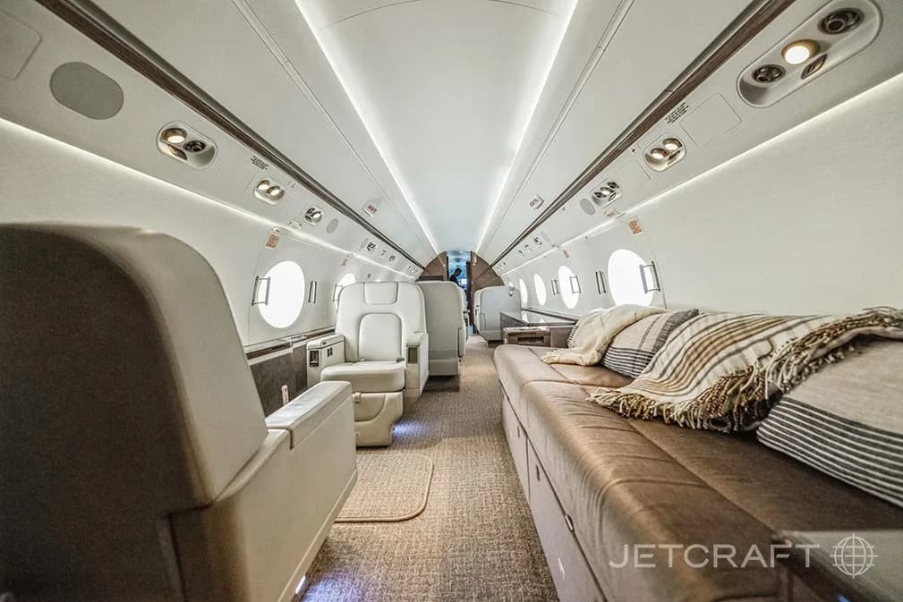 A long view of the aircraft cabin with a comfortable brown leather divan and individual light-colored seats.
