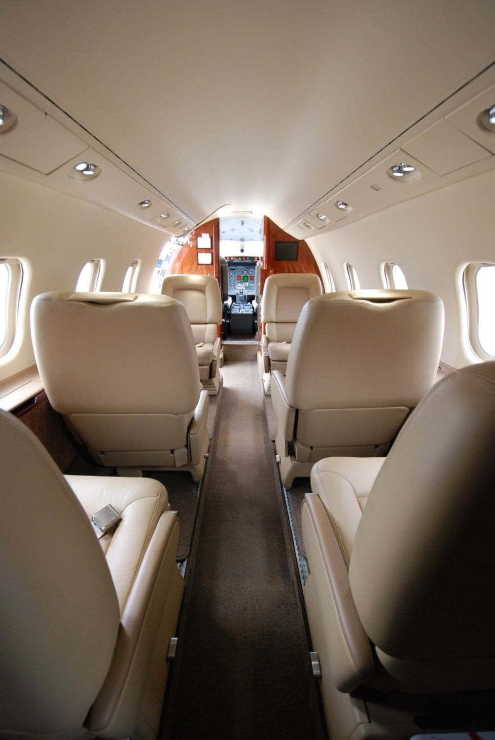 A view down the aisle of a business jet's cabin, showing beige leather seats and the cockpit entrance in the distance.