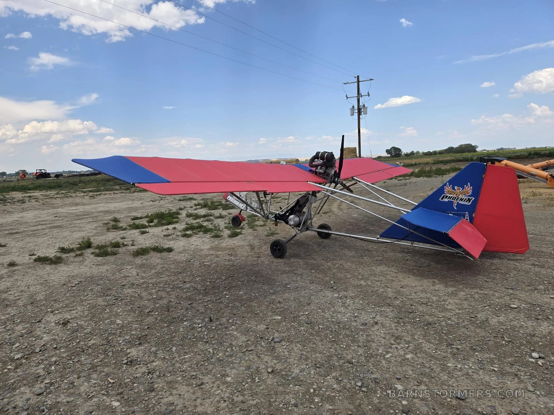 A red and blue ultralight aircraft with a 'Phoenix' logo on its tail, parked on a dirt field.