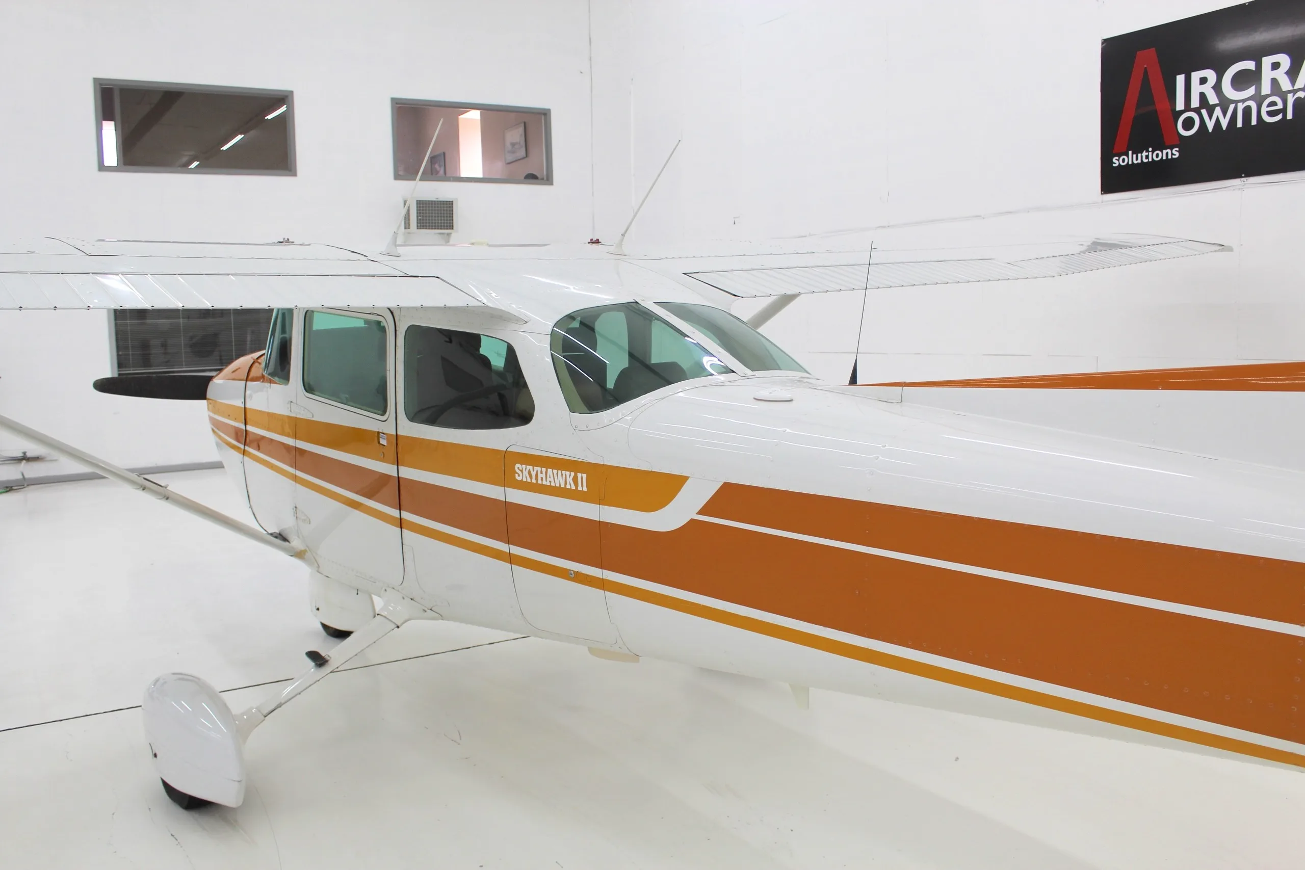 A white and orange Cessna Skyhawk II (N5546J) is parked in a brightly lit hangar, viewed from the front-side.