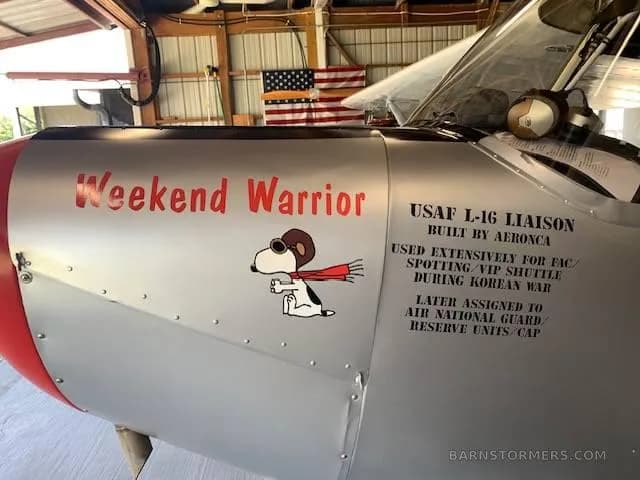 A close-up of the silver fuselage of the Aeronca L-16 aircraft, showing "Weekend Warrior" text, Snoopy nose art, and historical information.