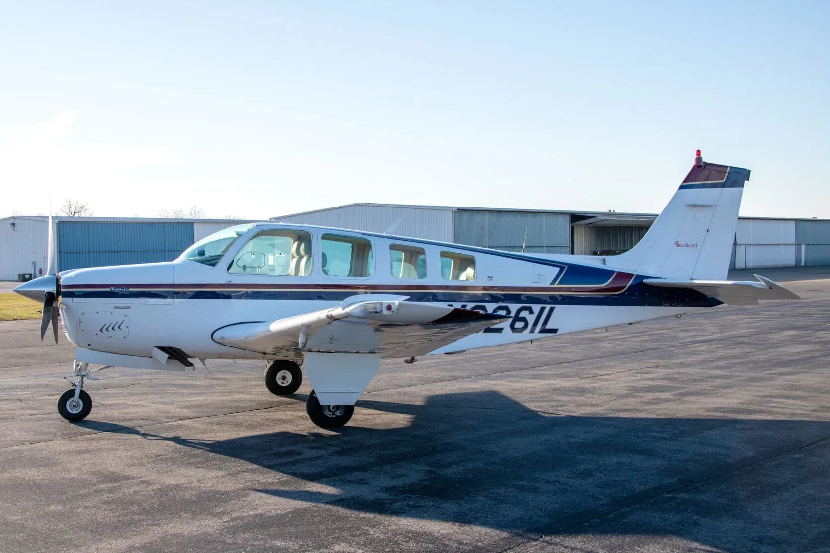 A white Beechcraft Bonanza N2261L with blue and dark blue stripes parked on an airfield.