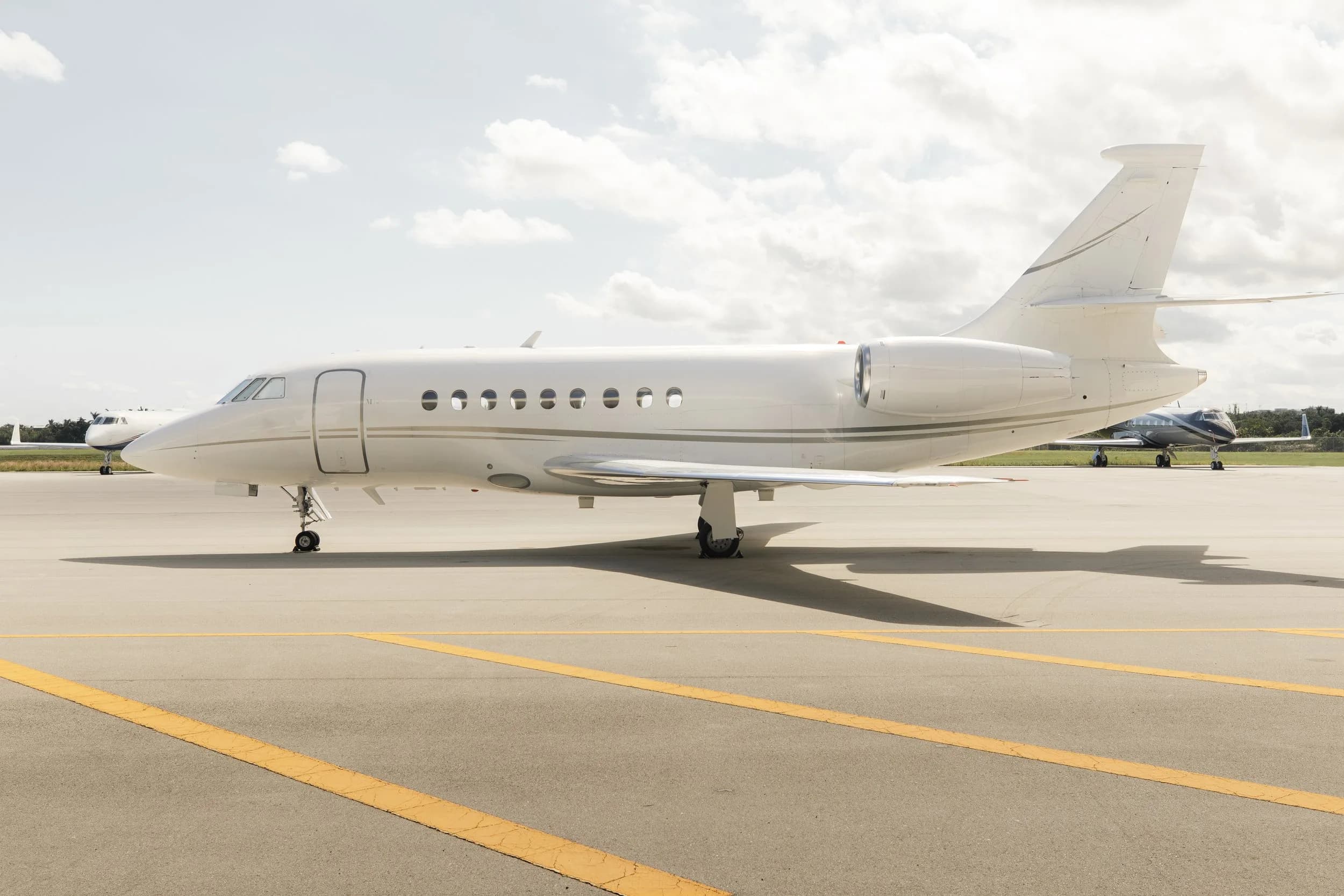 A white Dassault Falcon business jet with gold accents on the tarmac under a partly cloudy sky.