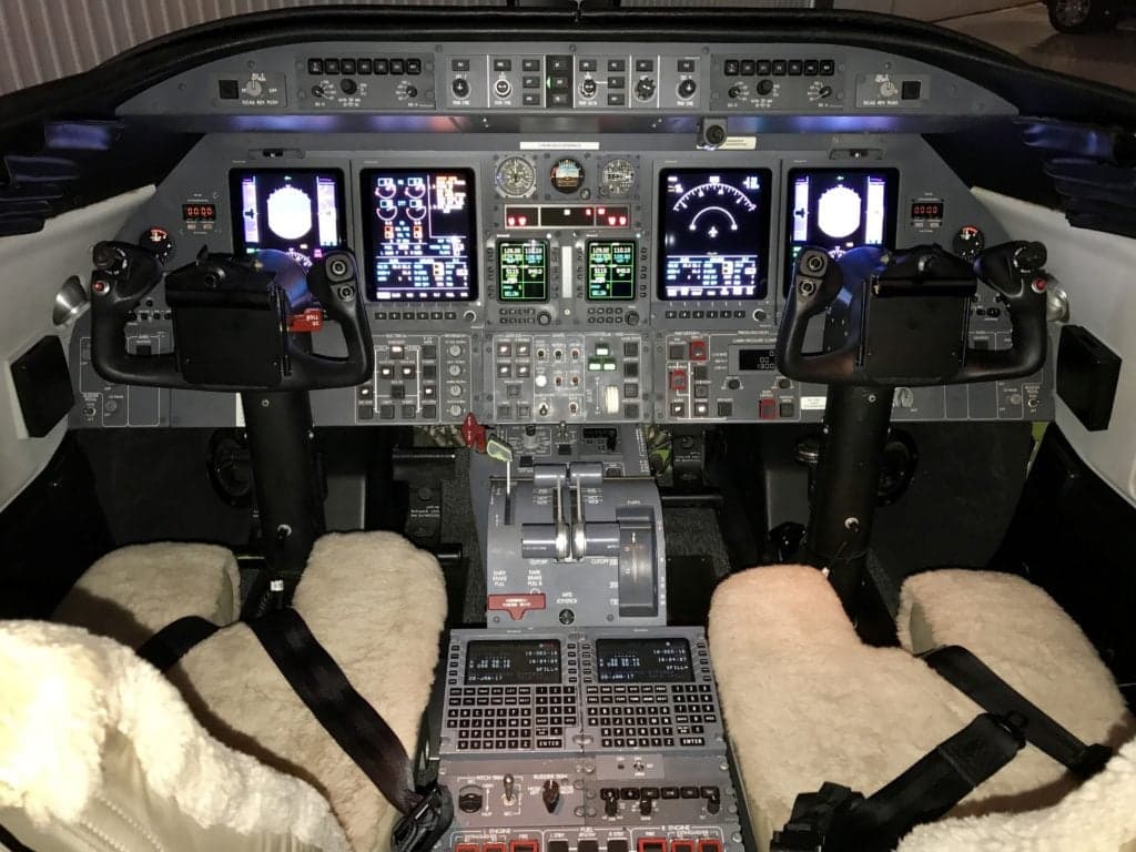A detailed view of a modern aircraft cockpit with multiple display screens, controls, and sheepskin seat covers.