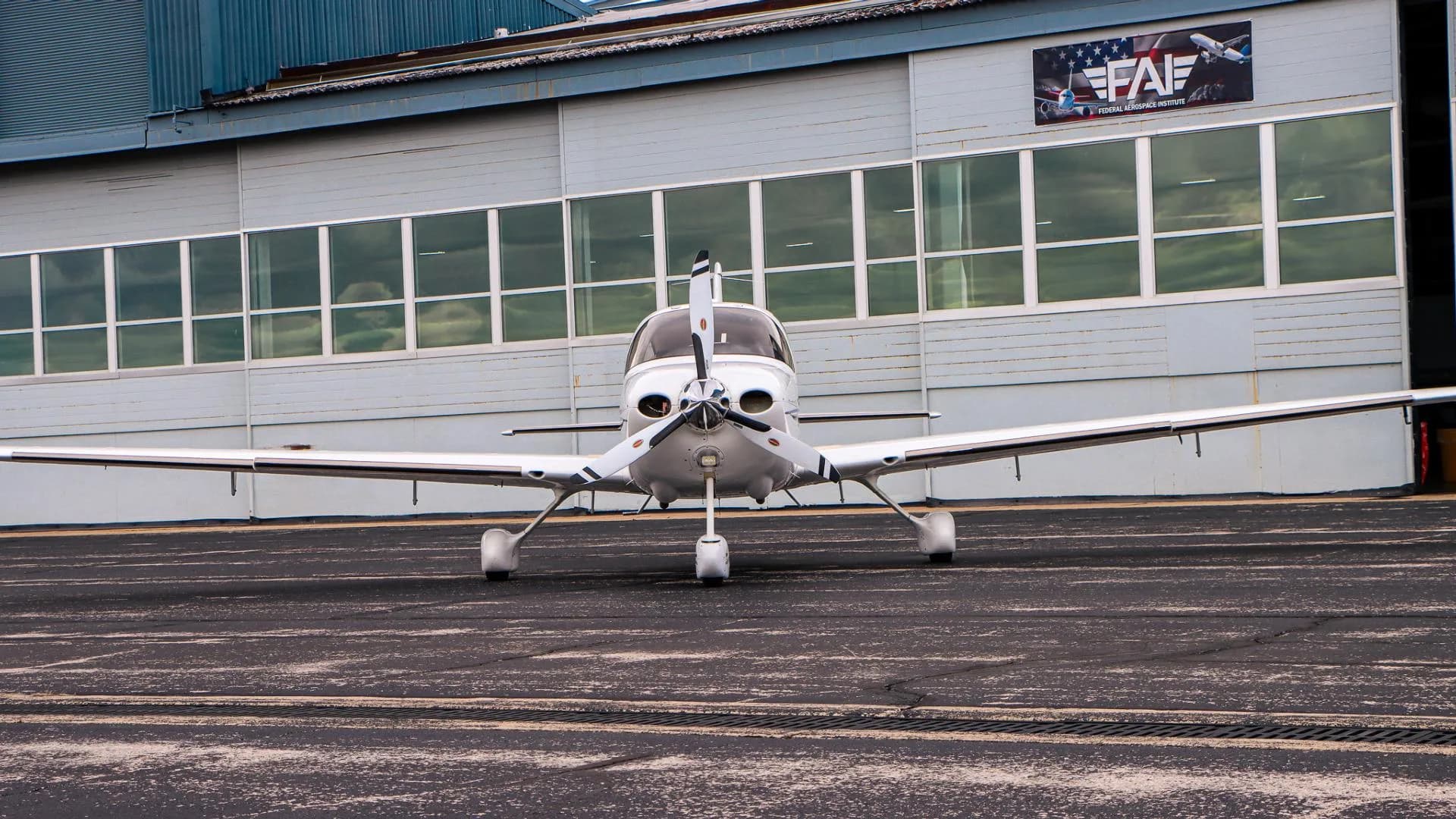 A white Cirrus aircraft with a three-bladed propeller is parked on a tarmac in front of a hangar.