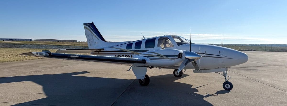 Front-left view of a white twin-engine propeller aircraft with black and gold stripes on an airfield.