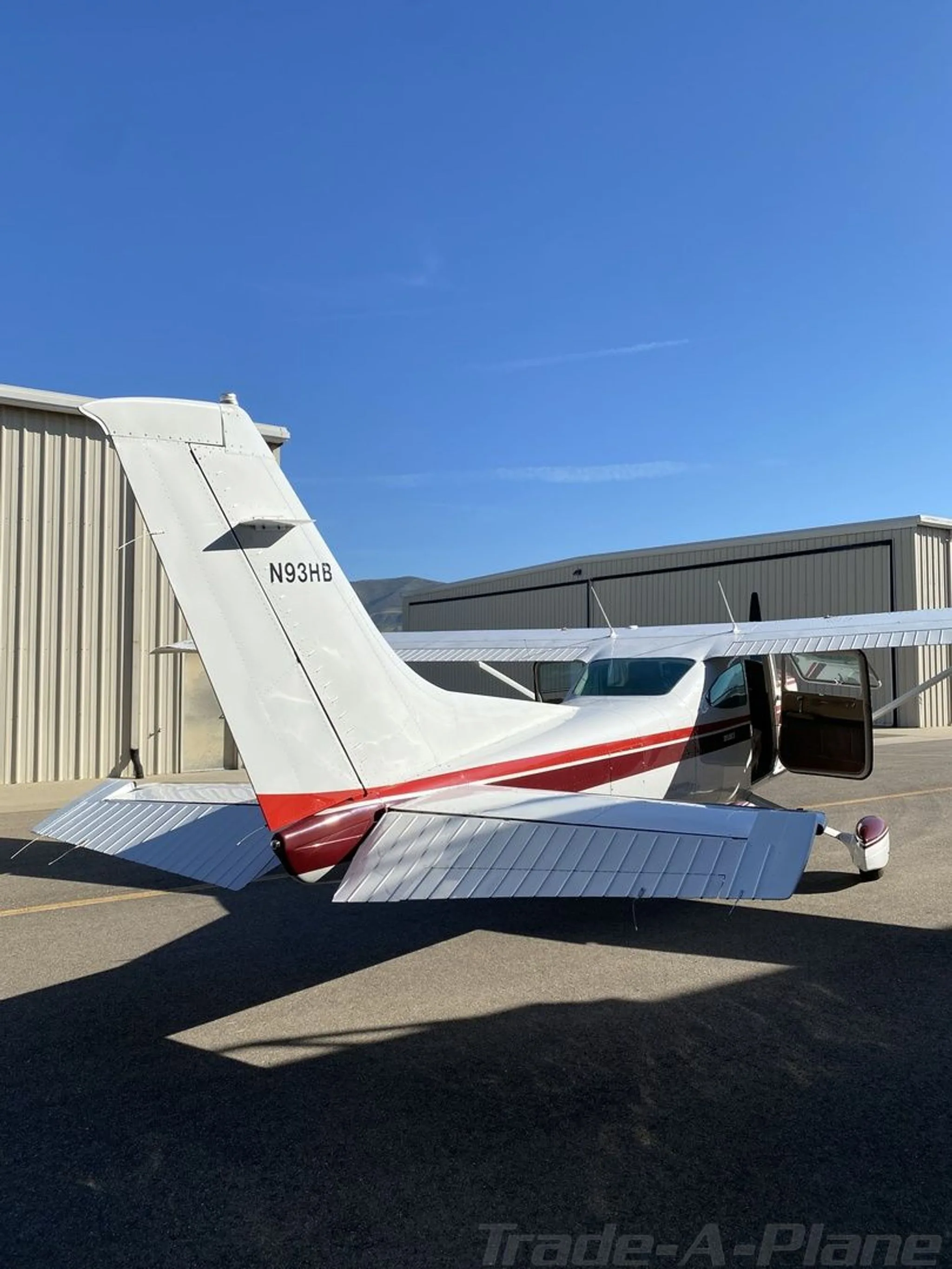A rear view of the white Cessna 182 with a red stripe, showing the open passenger door and N93HB registration on the tail.