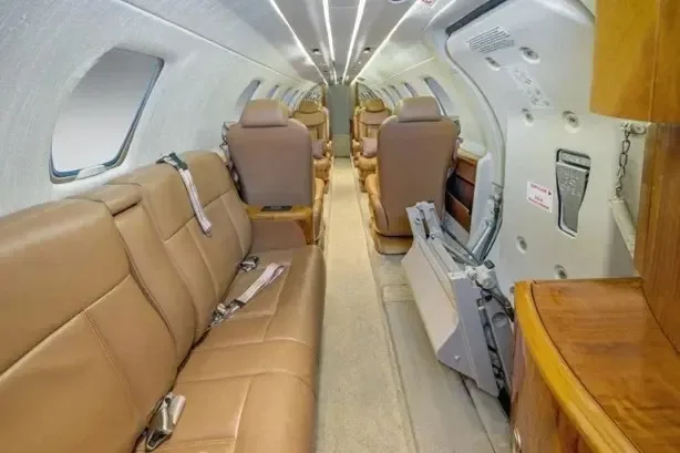 The aircraft cabin interior with a long tan leather couch on one side and individual seats on the other, looking towards the rear.
