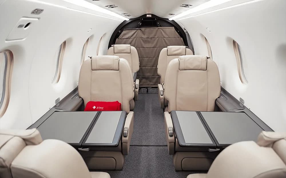 The interior cabin of a private aircraft, showing four beige leather seats with fold-out tables.