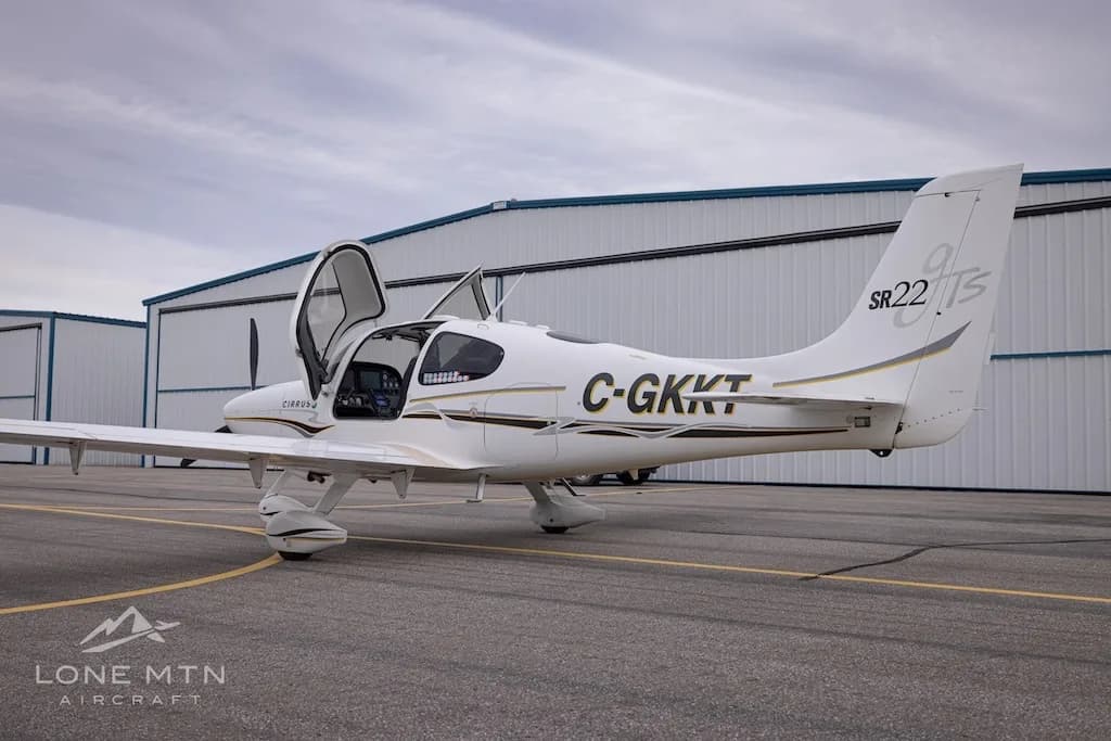 A slightly different rear three-quarter view of a white Cirrus SR22 aircraft with registration C-GKKT, doors open, on an airfield.