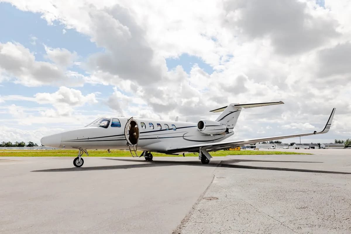 A white Cessna Citation business jet with its door open, parked on an airfield under a partly cloudy sky.