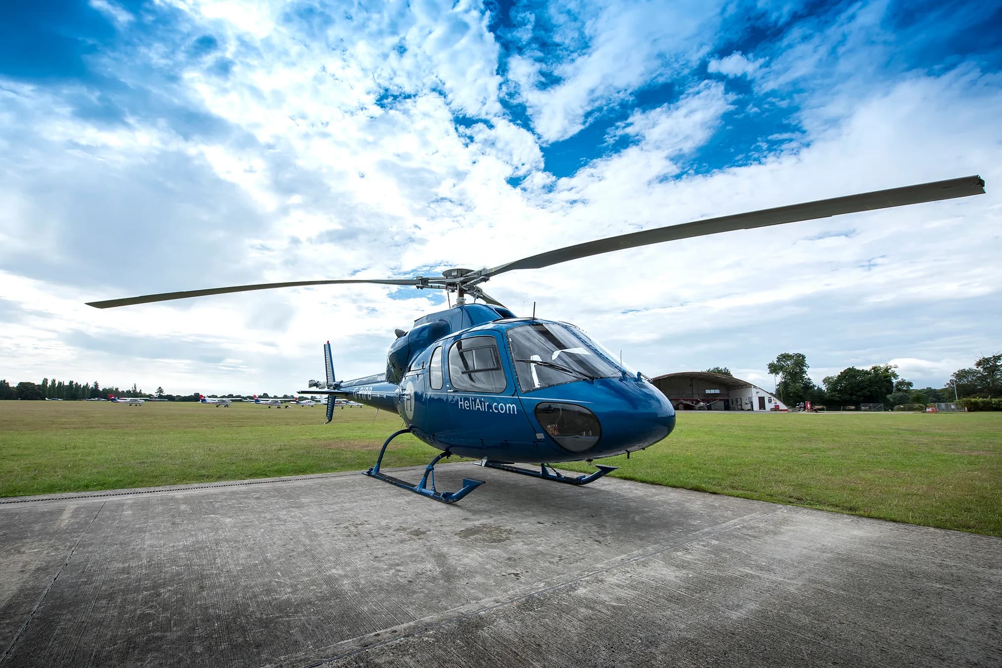 A blue helicopter with 'HeliAir.com' branding is parked on a concrete pad at an airfield under a cloudy sky.