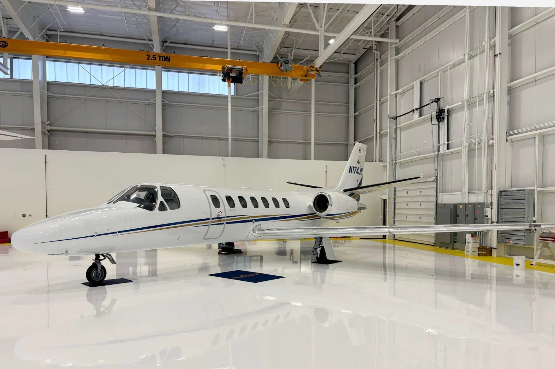 A white business jet with blue and gold stripes, tail number N174JS, parked inside a brightly lit hangar.