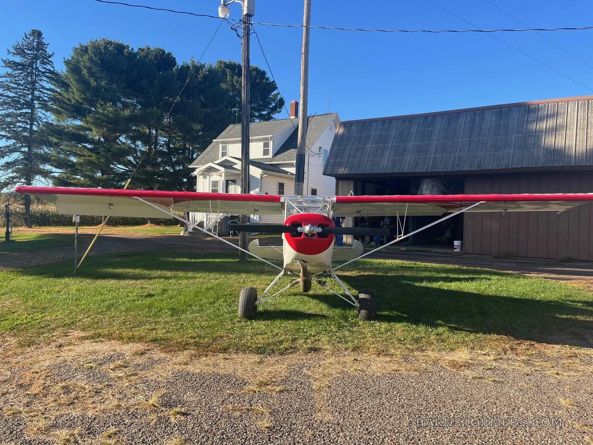 Front view of a white and red experimental aircraft with large tires, parked on grass and gravel in front of a barn and house.
