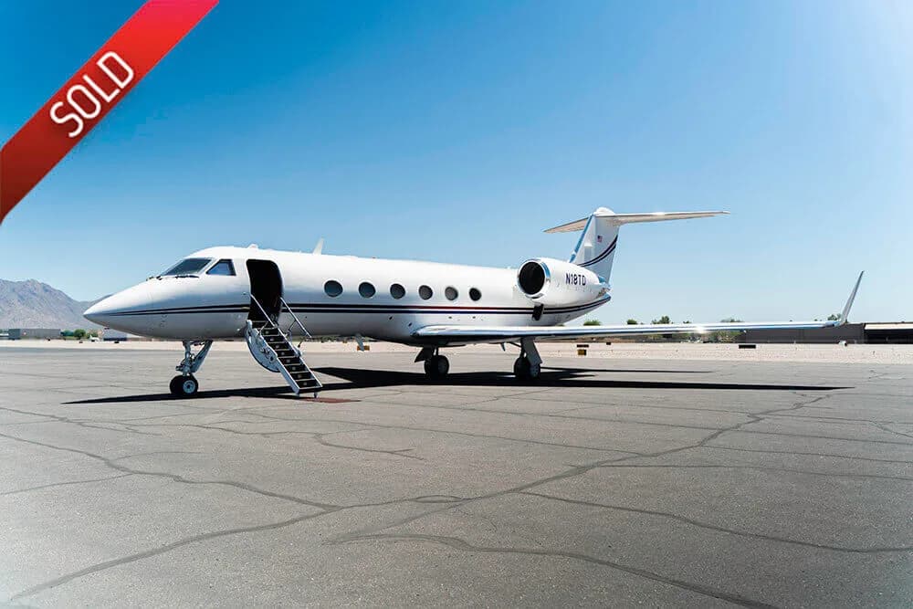 A white Gulfstream business jet with a blue and red stripe sits on an airport tarmac with its door open and stairs extended, under a clear blue sky.