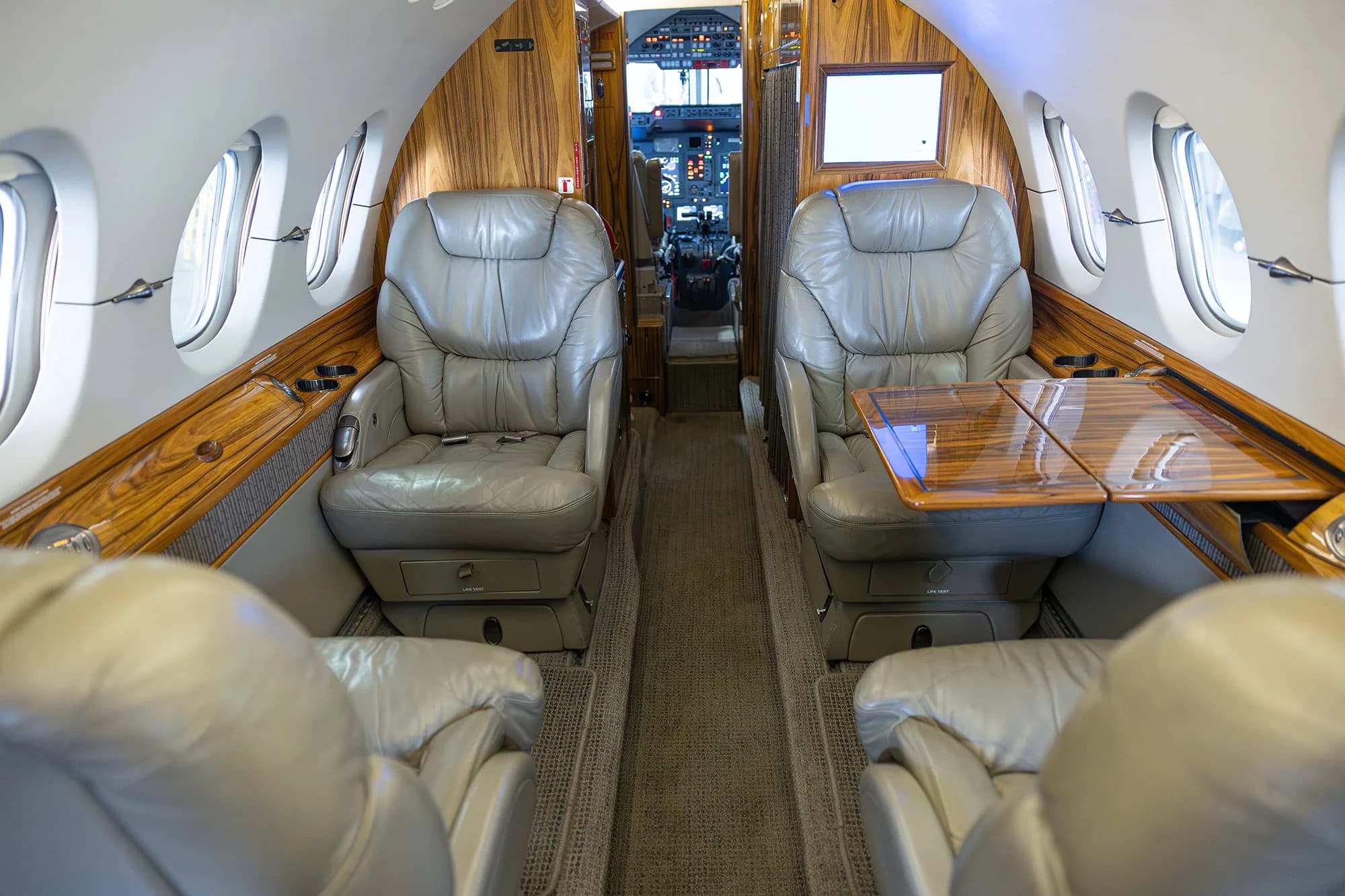 The interior cabin of a business jet with grey leather seats and wood paneling, showing a deployed table.