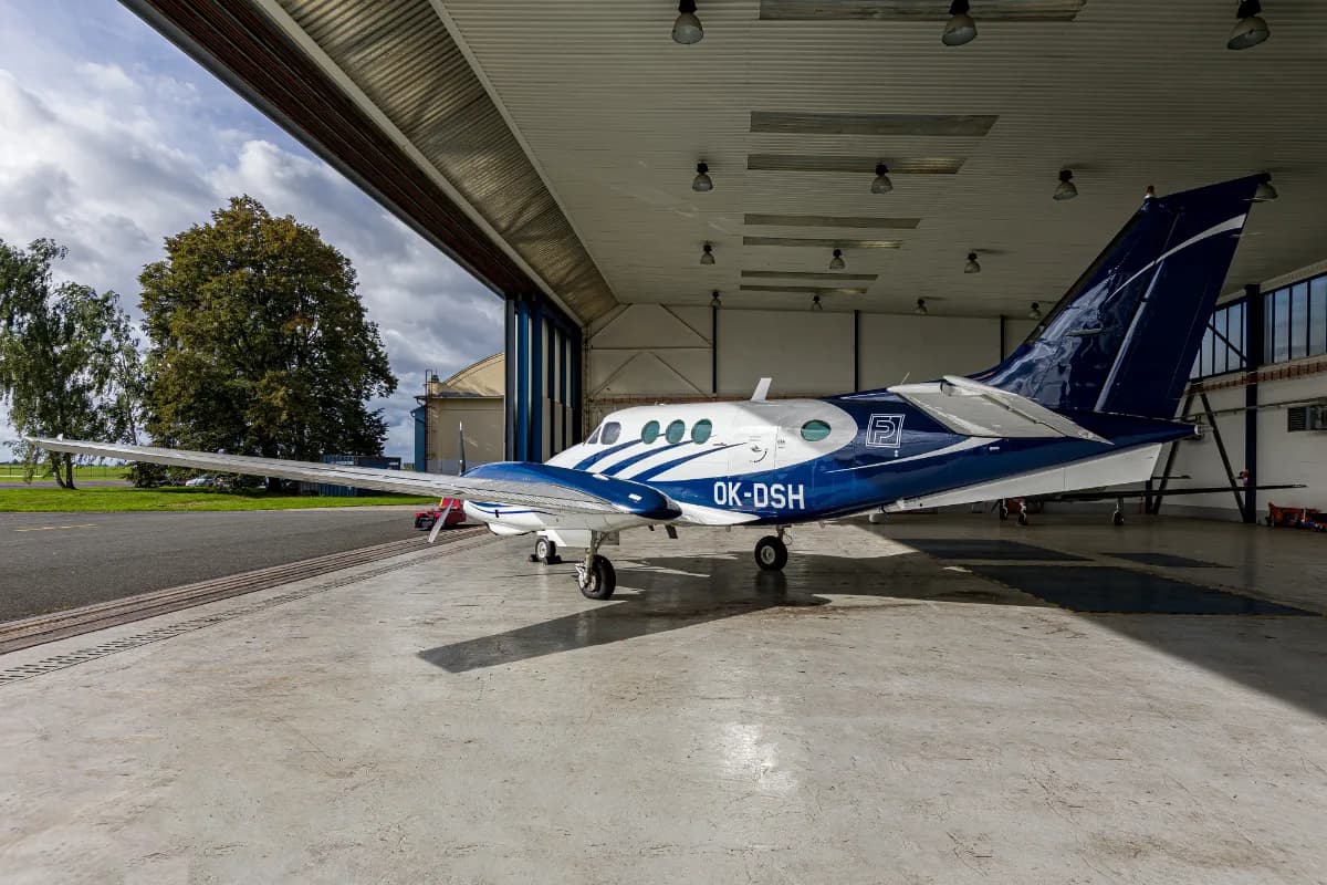 A white and blue Beechcraft King Air C90 twin-turboprop aircraft parked inside a hangar, with the hangar door open to an outdoor view.